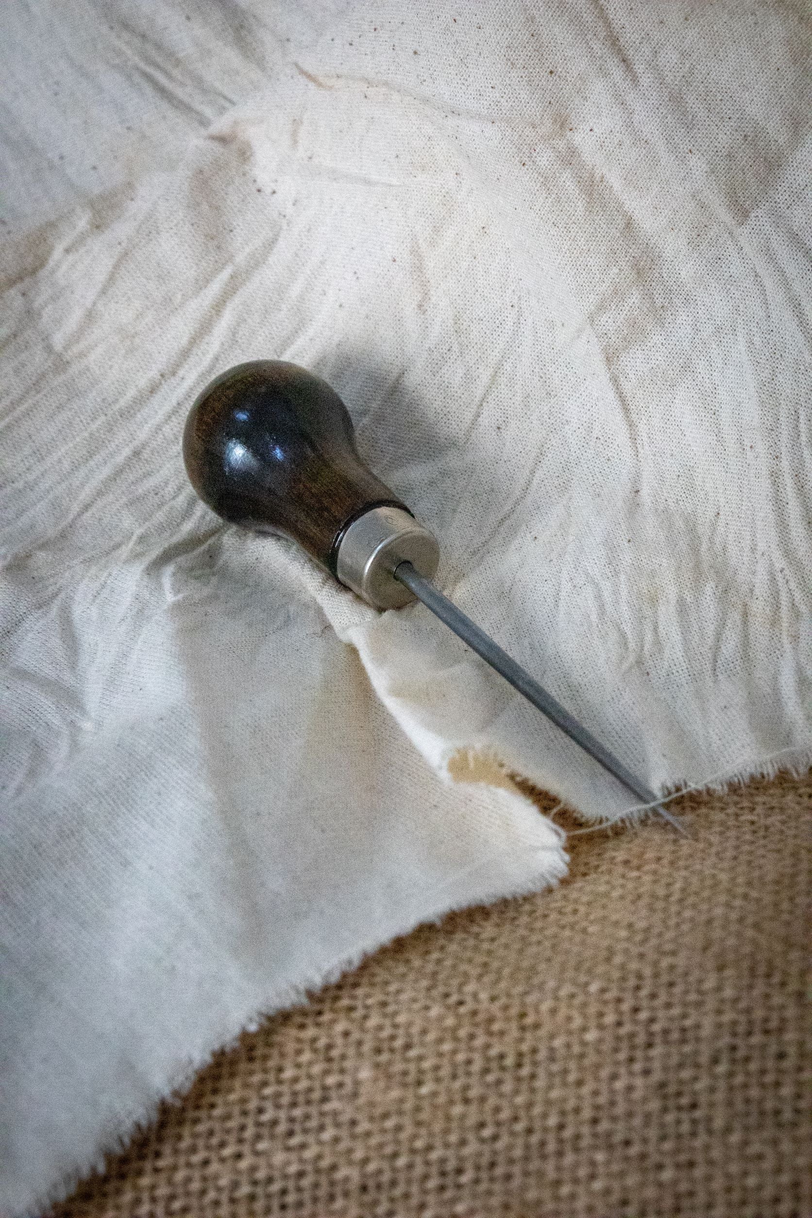 Full view of the RMIT Store Dart Marker Tailors Awl resting diagonally across layered cotton and hessian fabric, displaying the dark wooden handle and tapered steel shaft ideal for garment marking.